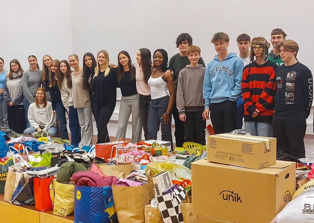 Eine Gruppe von Jugendlichen steht vor einem großen Stapel gespendeter Kleidung und Lebensmittel, die für eine Hilfsaktion gesammelt wurden.