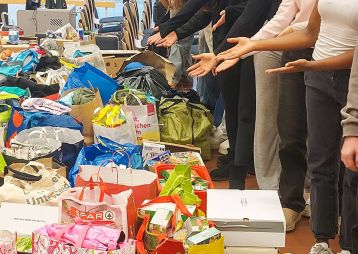 Eine Gruppe von Schülerinnen zeigt stolz auf die gesammelten Spenden in verschiedenen Taschen und Kisten, um Bedürftigen zu helfen.