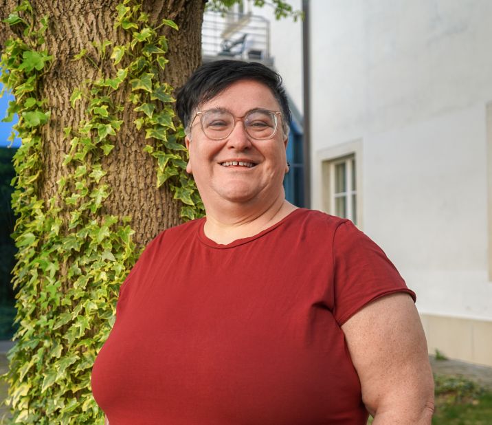 Eine Person mit Brille steht vor einem Baum, umgeben von Efeu, und lächelt freundlich.