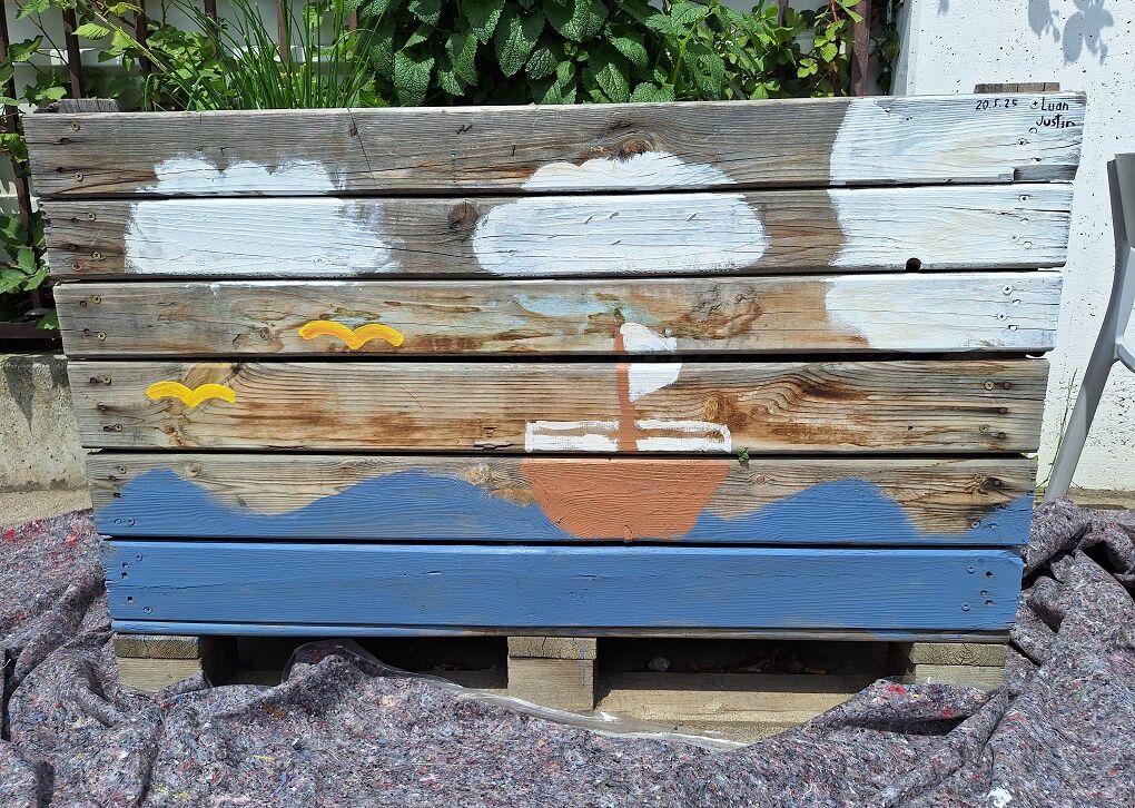 Ein Kunstwerk auf einem Holzbrett: Ein Schiff fährt über stilisierte Wellen, umgeben von Wolken und Vögeln.