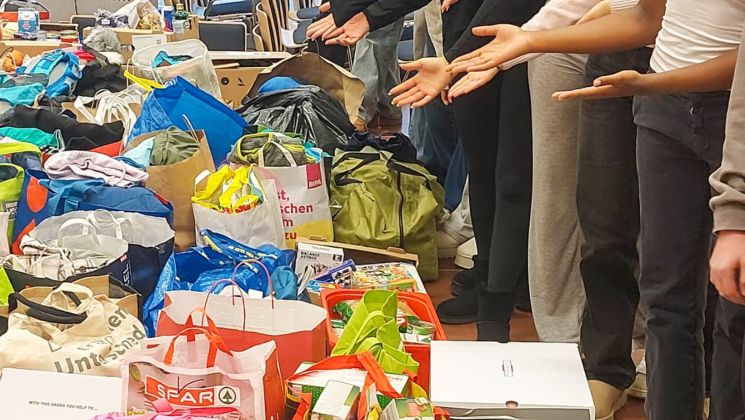 Eine Gruppe von Schülerinnen zeigt stolz auf die gesammelten Spenden in verschiedenen Taschen und Kisten, um Bedürftigen zu helfen.