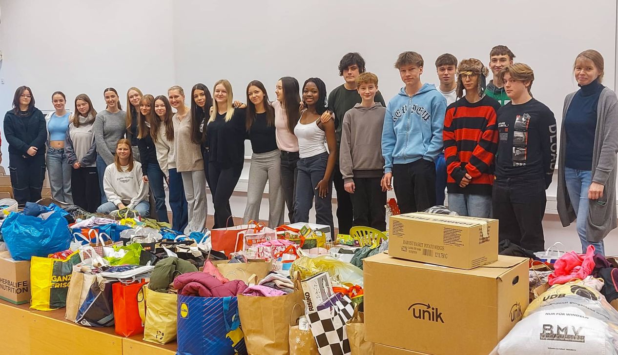 Eine Gruppe von Jugendlichen steht vor einem großen Stapel gespendeter Kleidung und Lebensmittel, die für eine Hilfsaktion gesammelt wurden.