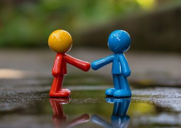 Zwei bunte Figuren, eine rote mit gelbem Kopf und eine blaue, schütteln sich die Hände auf einem nassen Untergrund.