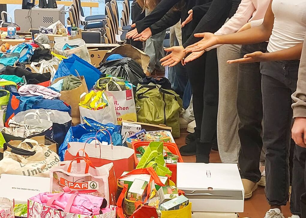 Eine Gruppe von Schülerinnen zeigt stolz auf die gesammelten Spenden in verschiedenen Taschen und Kisten, um Bedürftigen zu helfen.