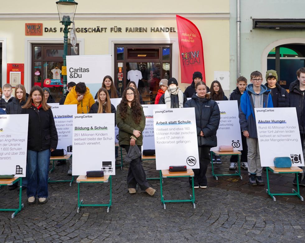 Das Bild zeigt Schüler*innen, die auf der Straße hinter Plakaten stehen. Sie machen aufmerksam auf Themen wie Bildung, Hunger, Schule und Kinderarbeit. Im Hintergrund sieht man ein Schild mit Caritas und Du.