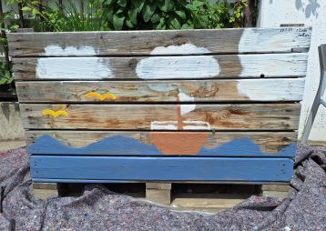 Ein Kunstwerk auf einem Holzbrett: Ein Schiff fährt über stilisierte Wellen, umgeben von Wolken und Vögeln.