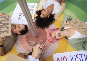 Vier lachende Menschen liegen in einem bunten Kreis und halten Schilder mit den Worten Frieden, Stolz und Gerechtigkeit.