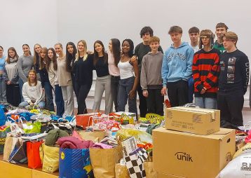 Eine Gruppe von Jugendlichen steht vor einem großen Stapel gespendeter Kleidung und Lebensmittel, die für eine Hilfsaktion gesammelt wurden.