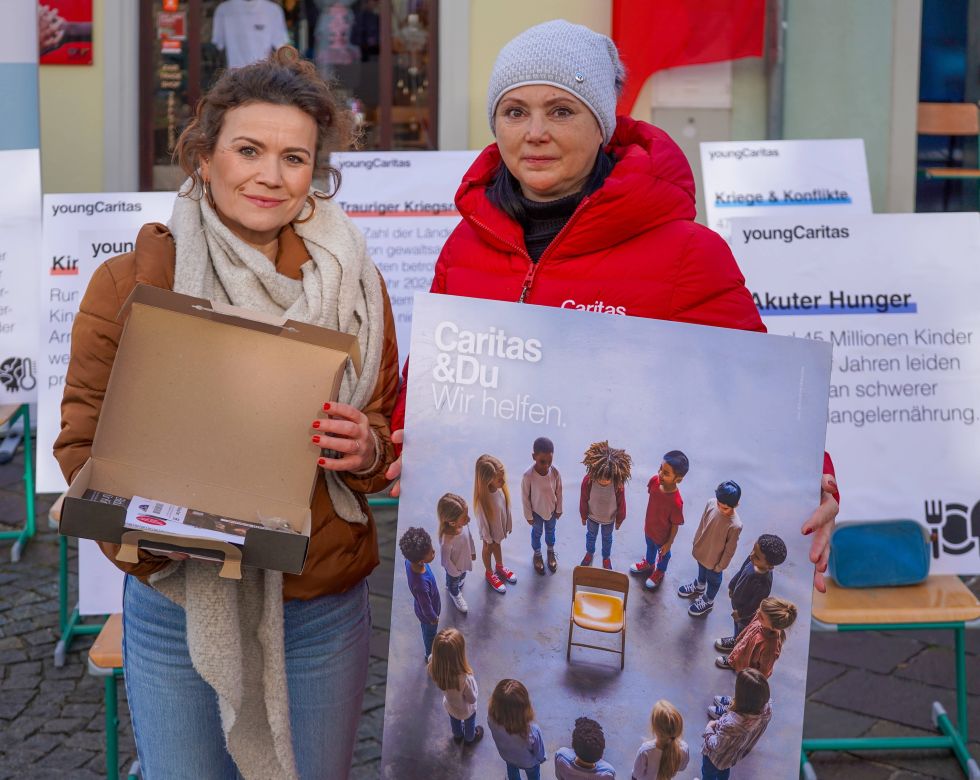 Zwei Frauen engagieren sich für Caritas, halten ein Plakat und eine leere Box, um auf Kinder in Not aufmerksam zu machen.