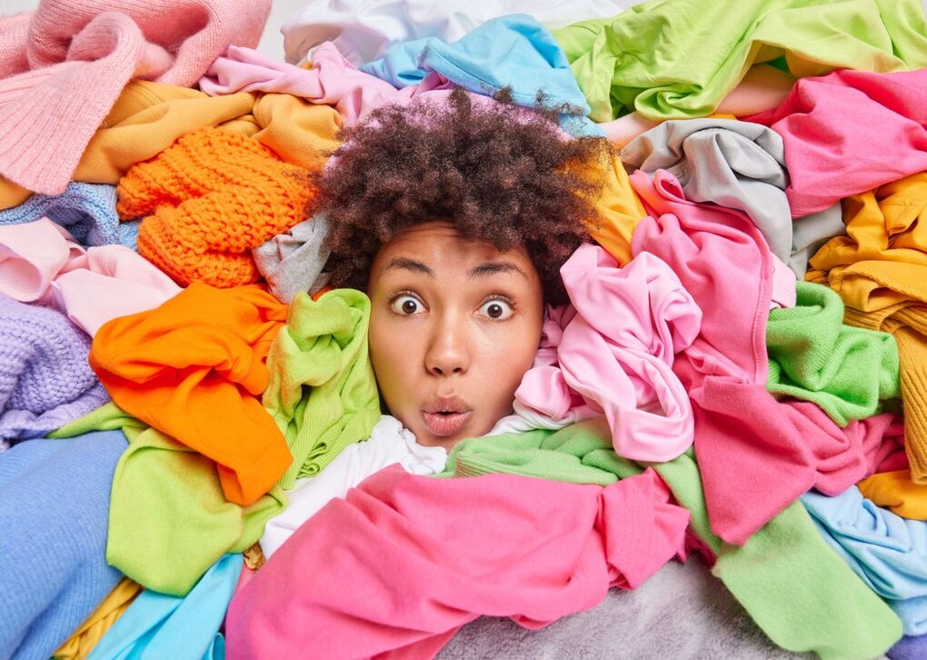 Eine Person ist von einem bunten Haufen Kleidung umgeben und sieht überrascht aus.