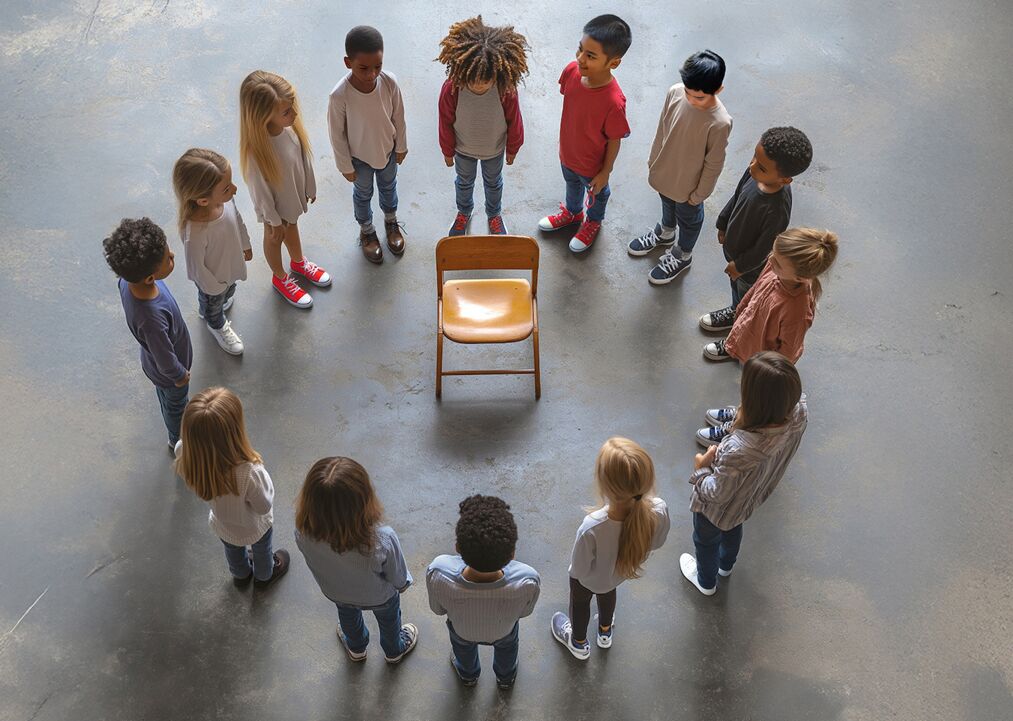 Eine Gruppe von Kindern steht im Kreis um einen Stuhl. Sie schauen auf den Stuhl, während sie in einem großen Raum stehen.
