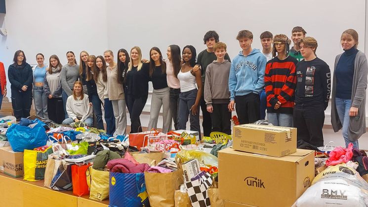 Eine Gruppe von Jugendlichen steht vor einem großen Stapel gespendeter Kleidung und Lebensmittel, die für eine Hilfsaktion gesammelt wurden.