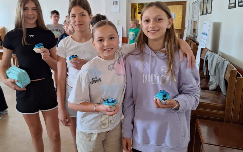 Vier Mädchen stehen nebeneinader in einem Raum und halten blaue Cupcakes in der Hand. Im Hintergrund sieht man Sesseln und weitere Kinder.
